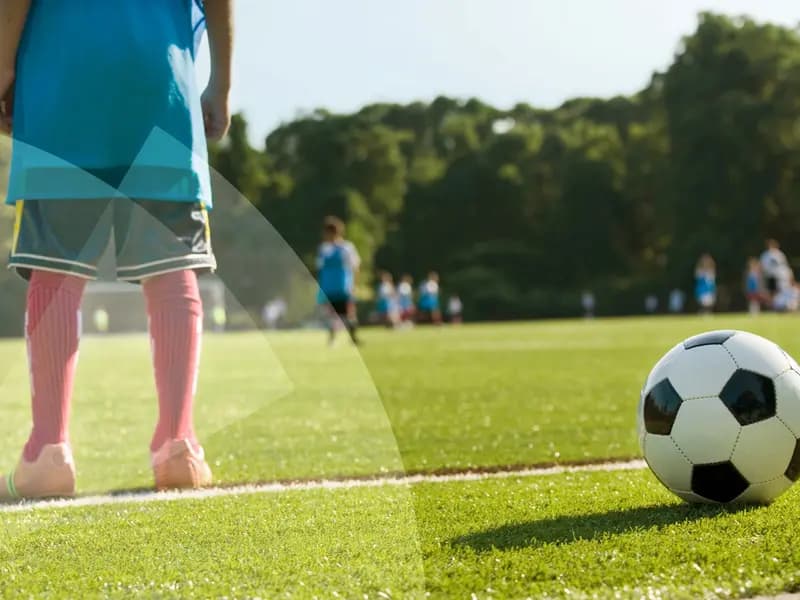 Kind im blauen Trikot neben einem Fußball auf einem Rasenplatz, im Hintergrund eine Jugendmannschaft