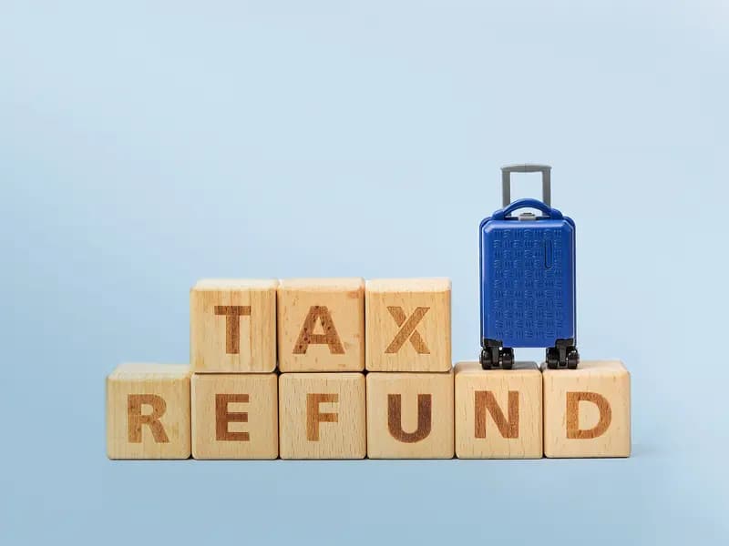 Holzwürfel mit der Aufschrift „TAX REFUND“ und ein kleiner blauer Rollkoffer
